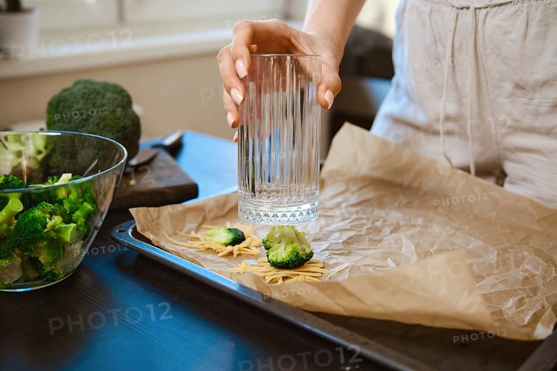 Woman is preparing smashed broccoli with cheese in a kitchen. She is using a glass to smash the broccoli on parchment paper. Fresh broccoli is nearby, showing the steps for a simple dish