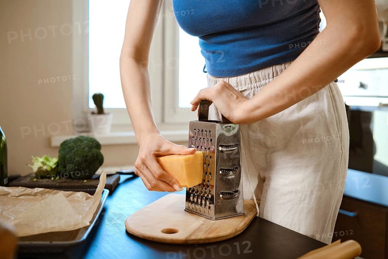 A woman grates cheese over a cutting board while preparing smashed broccoli in a kitchen. The bright sunlight comes through the window. Broccoli and other ingredients are nearby