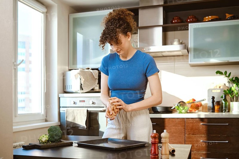 A woman crumples parchment to carefully lay it out on a baking sheet. She stands at a table, pressing the parchment in hands. The kitchen has various cooking tools and fresh ingredients around her