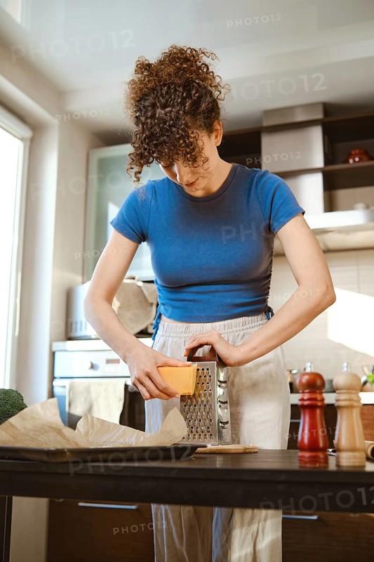 Woman grates cheese in a bright kitchen while preparing smashed broccoli. The scene shows cooking tools on the counter and the person focused on their task