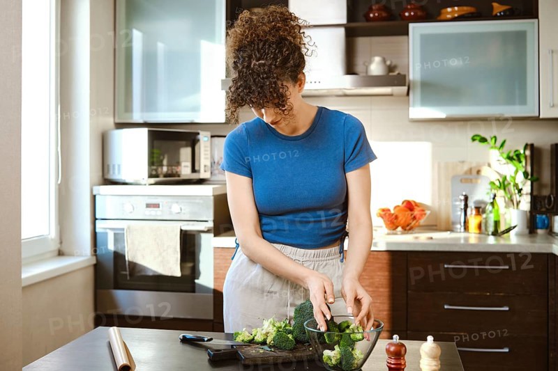 Woman shifting broccoli from chopping board into a glass bowl in a kitchen. The kitchen has sunlight coming in through a window