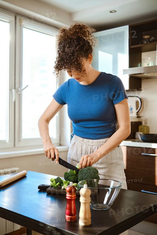 Woman chops broccoli in a kitchen. The cutting board is set on a table beside a bowl. Seasoning containers are nearby. Sunlight comes through the window, brightening the space