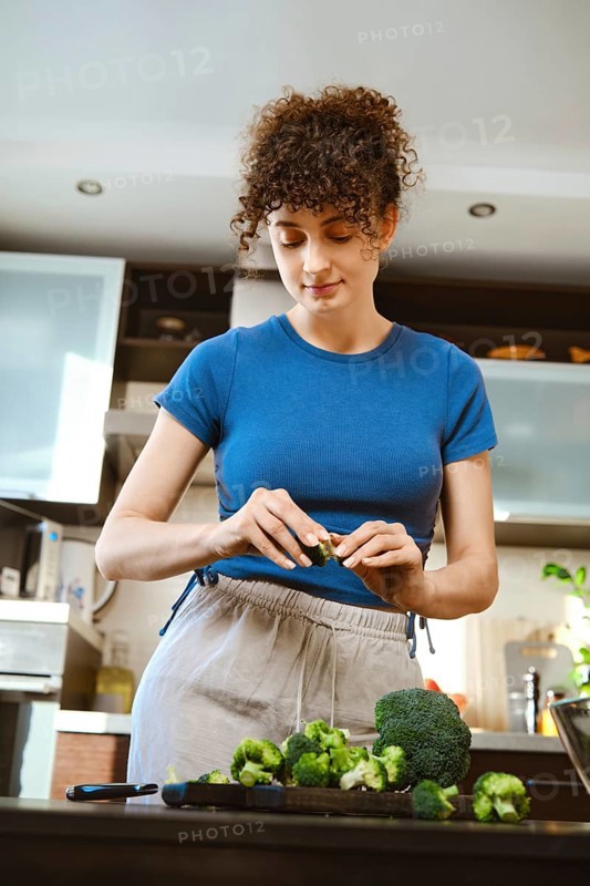 Woman is tearing up broccoli with fingers in a kitchen. The countertop has fresh broccoli. The person is focused on preparing the dish. The kitchen is well-lit and clean
