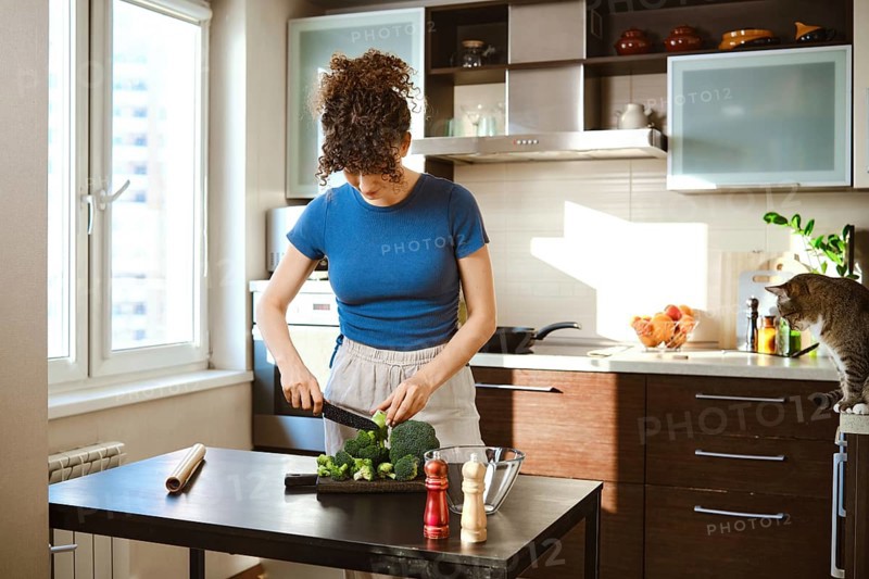 Woman is chopping broccoli on a black cutting board in a bright kitchen. The window shows daylight and a cat sits on the counter. Ingredients for cheese add flavor to the meal