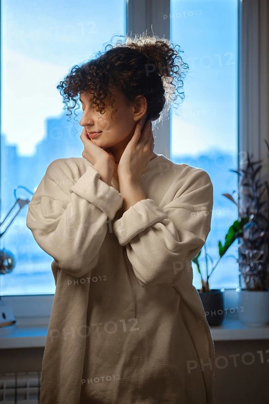 Young woman looks out the window while touching her face in a cozy indoor setting during daytime