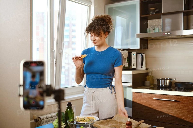 A food blogger records a video of cooking smashed broccoli with cheese. She stands in a bright kitchen and holds a piece of food in her hand