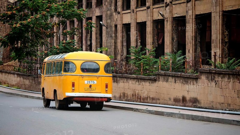 Old yellow bus runs on empty city street next to a weathered building, Alaverdi, Armenia