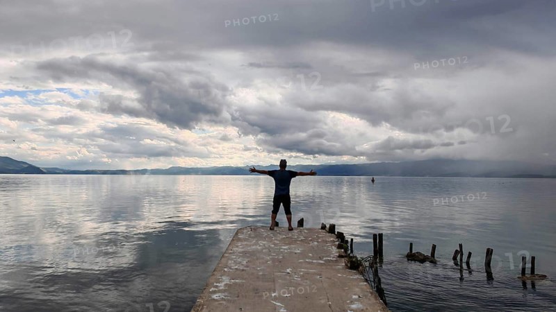 Person standing with outstretched arms on a pier surrounded by water and clouds, Lake Ohrid, North Macedonia