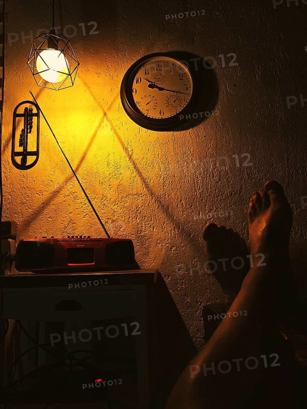 Cosy, warm scene with lamp and clock on the wall, cast shadows and a relaxed leg on the table