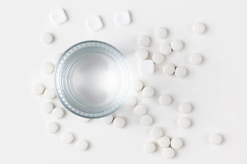 Glass of water with lots of scattered white tablets on a white surface