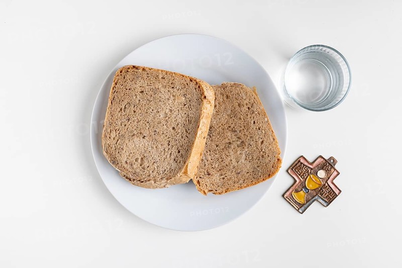 Two slices of bread on a plate with water glass and cross symbol on a plain white background, Lent