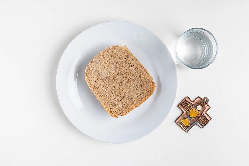 Slice of bread on a plate with a water glass and a cross symbol on a plain white background, fasting