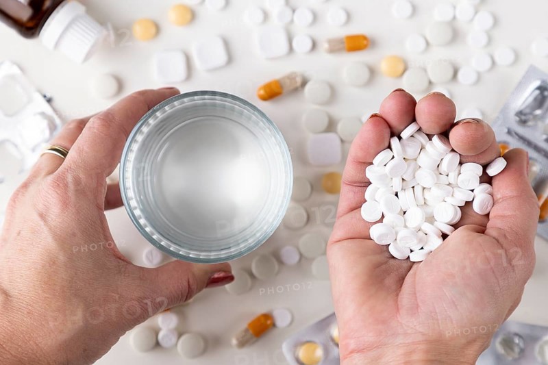 Two hands hold a glass of water and a handful of tablets across a table