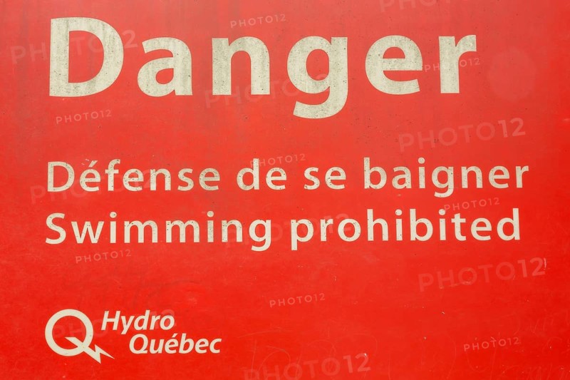 Bilingual French and English Danger Swimming Prohibited in white letters on red painted metal public notice sign posted by Hydro-Quebec utility in public park, Quebec, Canada