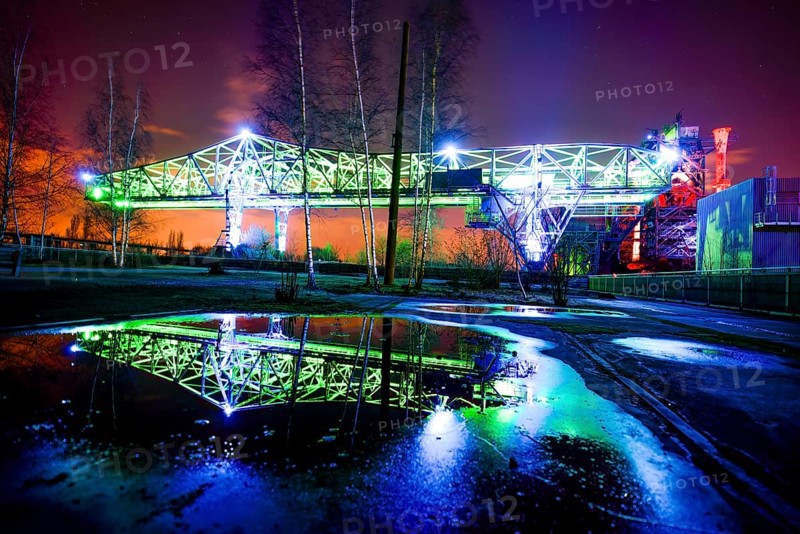 Illuminated bridge at night with blue and green lighting reflecting in a pool of water, Landschaftspark Duisburg Nord, Duisburg, North Rhine-Westphalia, Germany