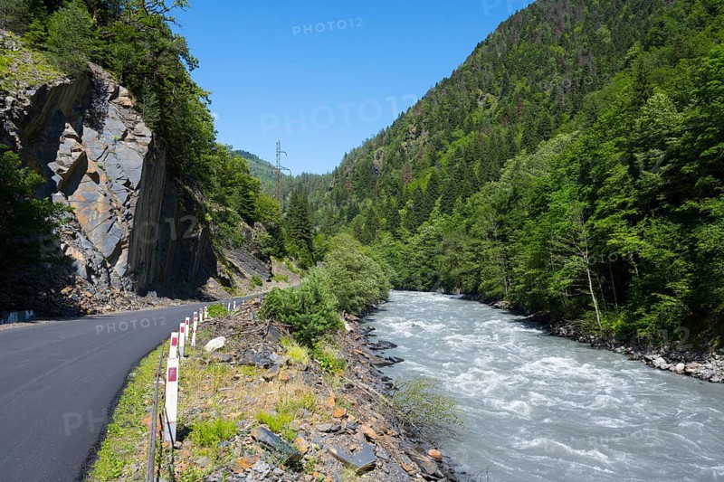 Mountain road next to a flowing river in a green, wooded landscape in clear summer weather, Enguri river between Khaishi and Tsalanari, Mingrelia region and Upper Svaneti, Georgia