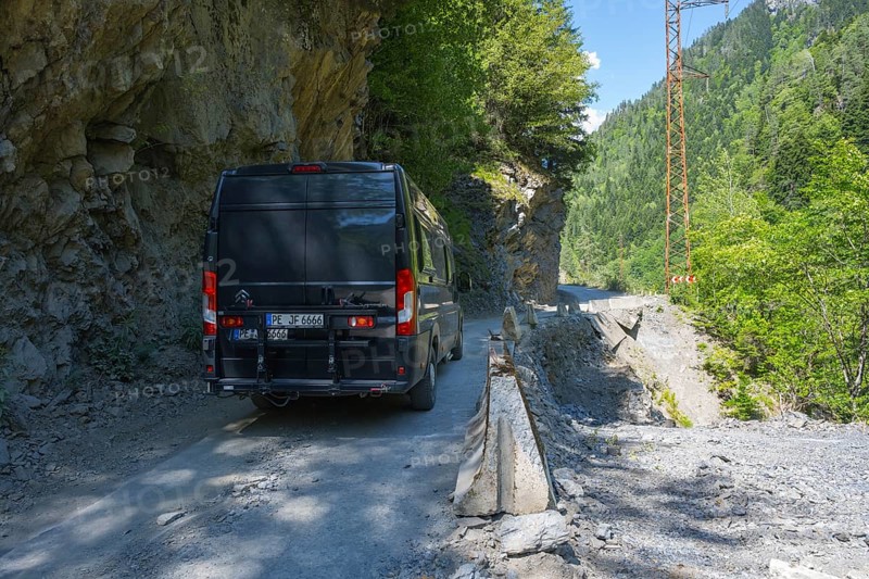 A black van drives on a narrow mountain road surrounded by rocks and thick vegetation, camper drives on destroyed road 7 between Khaishi and Tsalanari, Mingrelia region and Upper Svaneti, Georgia