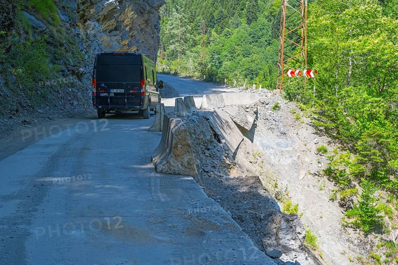 A black van drives on a remote road along a steep cliff with green vegetation, camper drives on destroyed road 7 between Khaishi and Tsalanari, Mingrelia region and Upper Svaneti, Georgia
