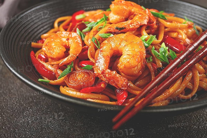 Udon stir-fry shrimp noodles with red bell pepper and green onion, served in a black bowl, with chopsticks, dark background