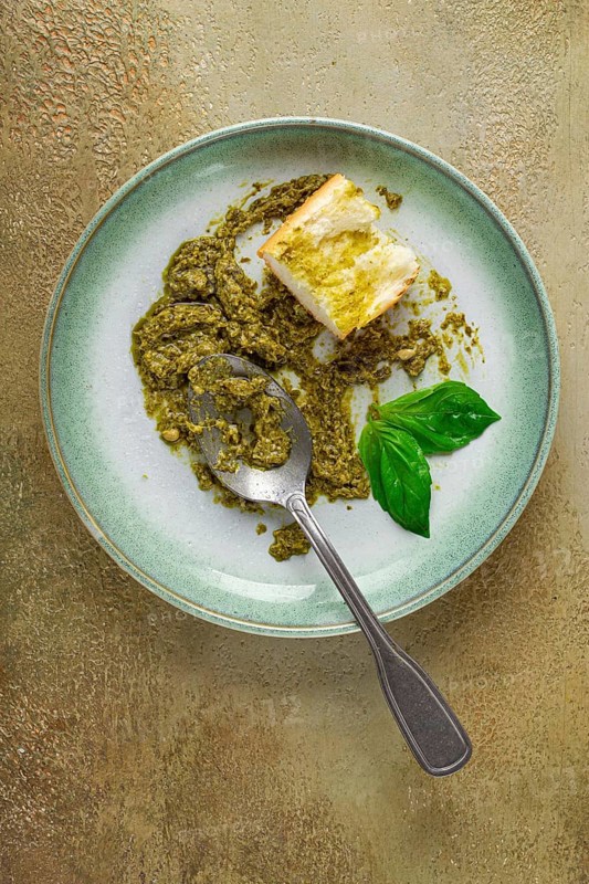 On a platter is a pesto sauce made from fresh basil, with a slice of bread and a spoon on a plate. The pesto sauce looks creamy and green, and the bread is partially eaten