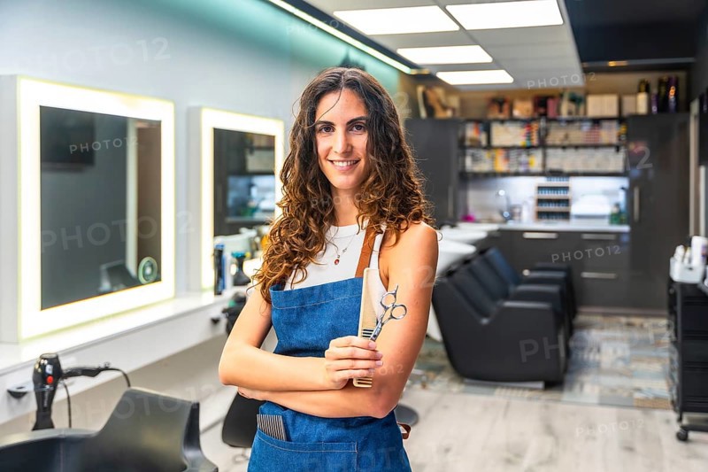 Professional female hairdresser in a modern salon smiling confidently while holding scissors and a comb, standing in apron ready to style hair and welcome clients