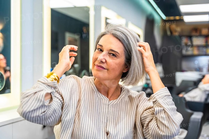 Senior woman smiling and touching her healthy gray hair, sitting in a modern hair salon, ready for a new hairstyle, embracing natural beauty and self care