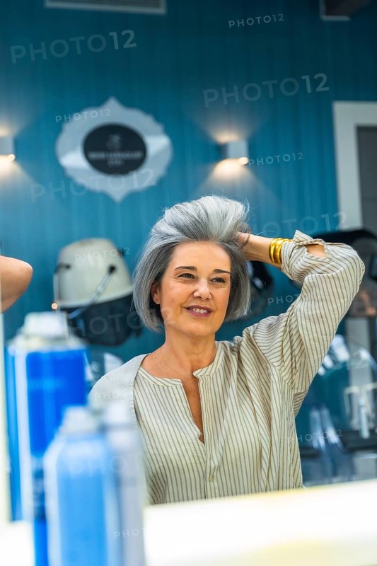 Mature woman looking at her reflection, touching her fresh gray hairstyle, and smiling in a modern beauty hair salon, feeling confident and happy with her new look