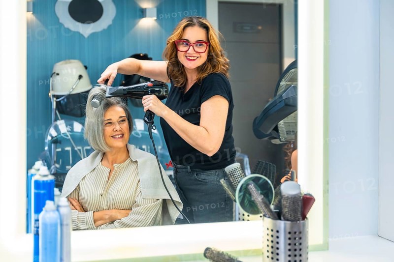 Female hairdresser smiling at camera while blow drying a mature customer's hair in a modern salon, creating a stylish look with professional tools and attentive service
