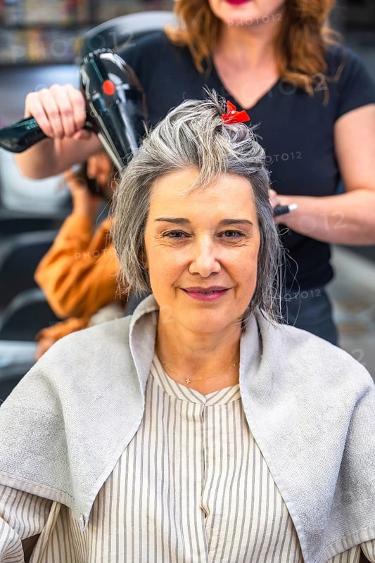 Mature woman sitting in a hair salon during a blow dry session, smiling confidently while a professional hairstylist works on her grey hair, providing care and beauty treatment