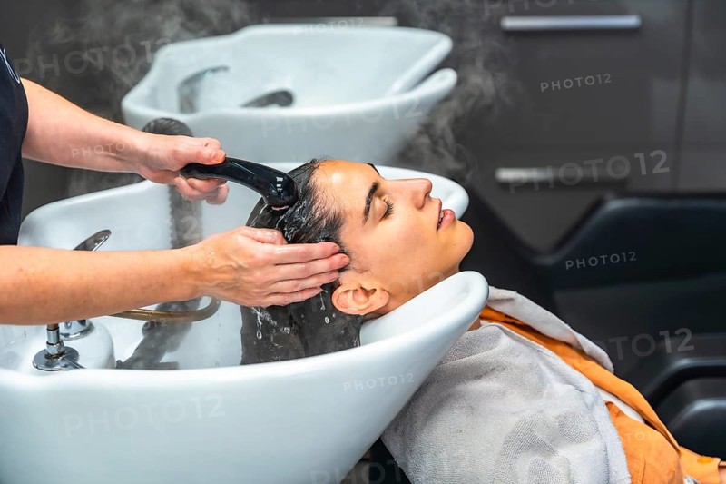 Woman relaxed in salon basin as hairstylist rinses shampoo and massages scalp with warm water and foam, creating a tranquil, pampering hair treatment moment