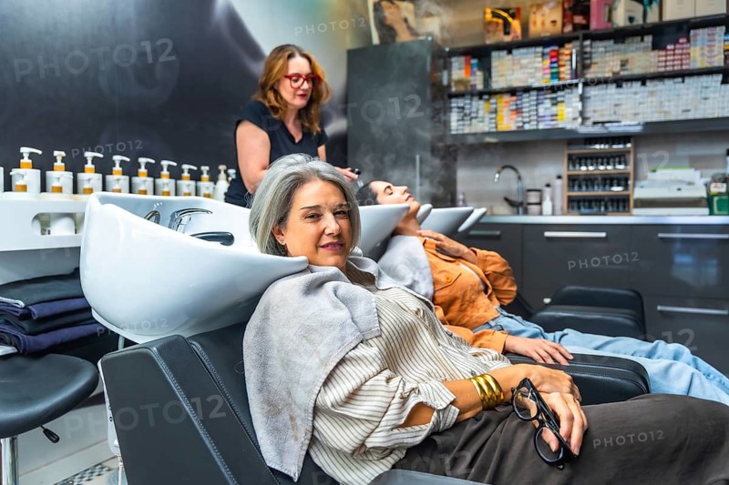 Two women reclining at salon basins while a stylist performs professional shampoo and scalp care, conveying relaxation, pampering, and modern haircare for diverse, mature clients