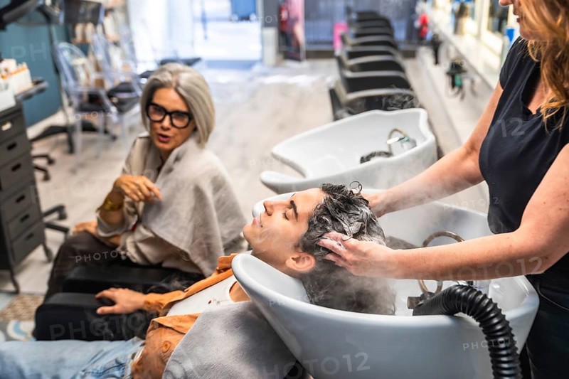 Professional hairdresser washing a young client's hair at the shampoo basin, providing hair care services in a contemporary hair salon while another customer waits