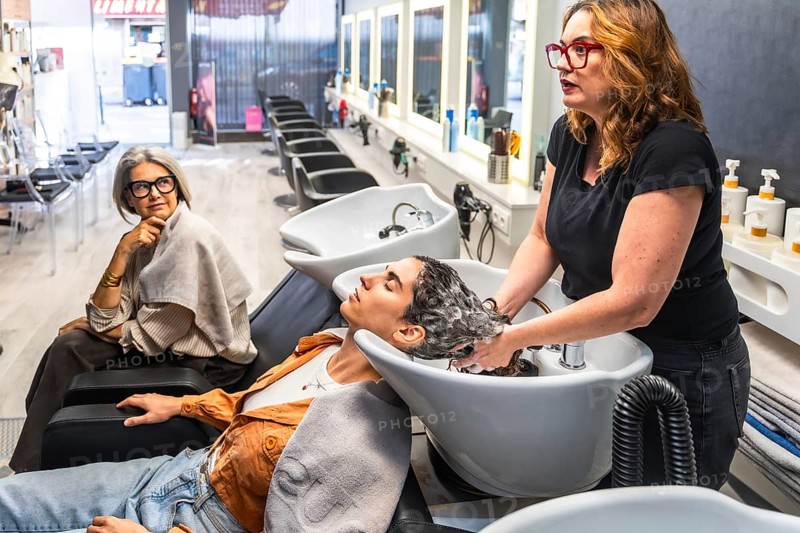 Hairdresser shampooing a young woman at a modern salon while an older client watches nearby, emphasizing professional hair care, pampering, comfort and clean, stylish interior