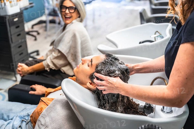 Hairdresser washing customer's hair at basin, providing relaxing care while another woman smiles happily in the background, a common scene in a modern beauty salon
