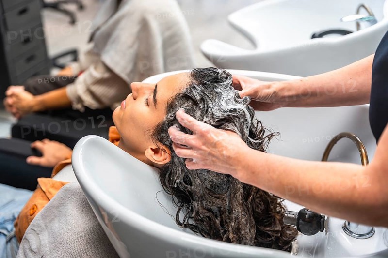 Woman feeling relaxed while a hairdresser is shampooing her long, curly hair with foam and water at a modern hair salon, providing a professional hair care treatment