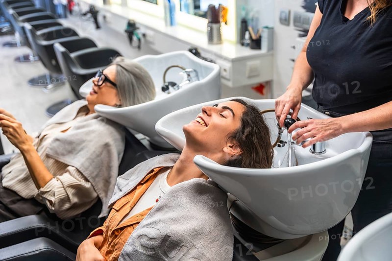 Female hairdresser washing a young woman's hair in a salon hair wash basin, while another client waits nearby, enjoying a relaxing moment during a beauty treatment
