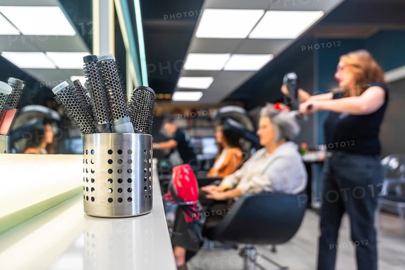 Hairbrushes standing ready on a white counter as a stylist blow dries a senior woman's hair in a modern hair salon, focusing on hair care and beauty services