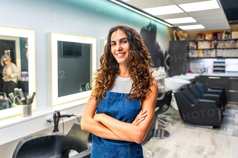 Confident young woman with curly hair wearing an apron and smiling, standing with crossed arms in her modern hair salon, representing a small business owner