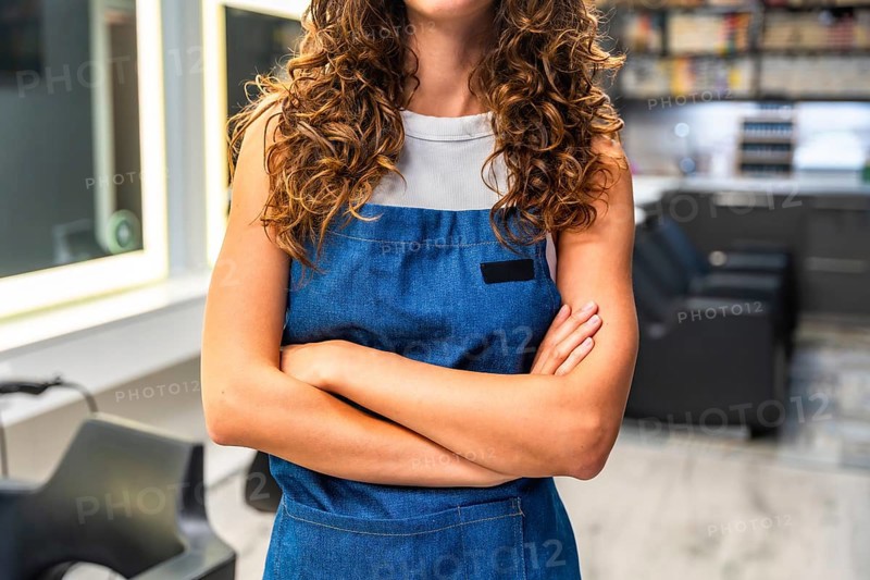 Professional hairdresser with curly hair standing confidently in a hair salon, wearing a stylish denim apron and crossing her arms, ready to start her work in the beauty industry