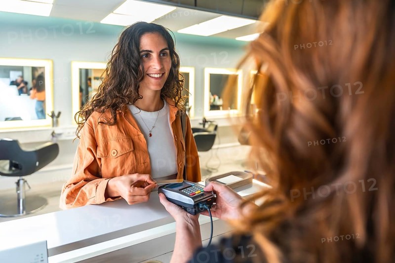 Smiling woman paying with a contactless credit card at a modern hair salon reception, completing a beauty service transaction at the checkout counter with ease and satisfaction
