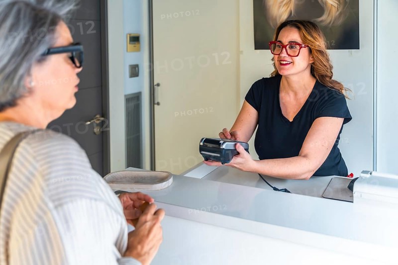 Salon receptionist smiling and processing a contactless credit card payment at the front desk for a mature woman customer with grey hair and glasses, friendly transaction