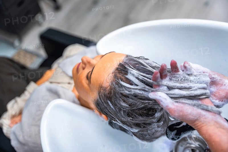 Mature woman relaxing with eyes closed as a hairdresser shampoos her long dark hair over a salon wash basin, creating rich suds and a soothing head massage