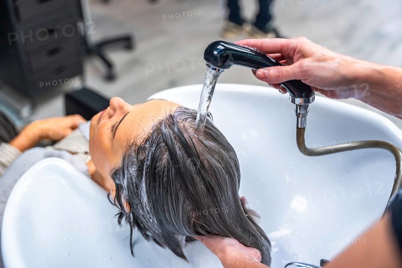 Woman relaxing as hairdresser rinses her hair over a salon sink with a handheld shower, receiving professional shampoo treatment and pampering care for clean, healthy hair