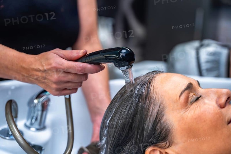 Hairdresser's hands rinsing a woman's hair with water from a shower head at a salon basin, focusing on hair care and relaxation during a beauty treatment