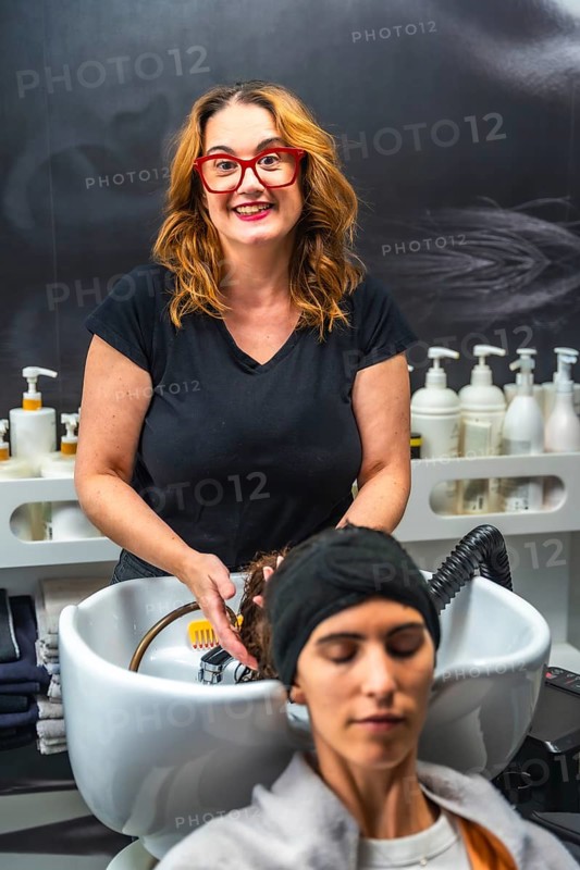 Hairdresser smiling while washing client's long red hair over salon sink, providing professional shampoo and relaxing hair care treatment in a bright, clean salon setting