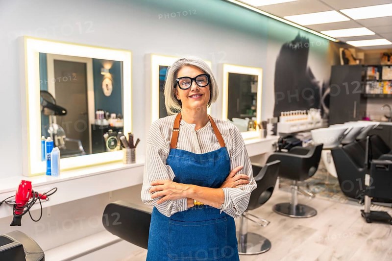 Senior woman hairstylist smiling, standing with arms crossed, wearing glasses and an apron, confidently managing her hair salon storefront, representing entrepreneurship and small business success