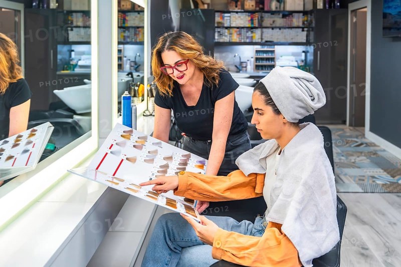 Hairdresser consulting with client on hair color selection at a modern salon, with the client pointing at shades on a color swatch chart while wearing a towel after hair wash