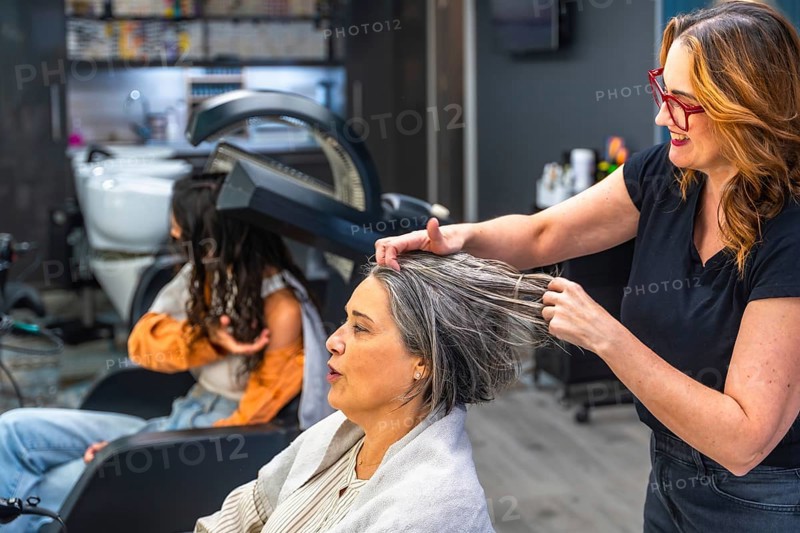 Hair stylist applying product to a senior woman's hair in a professional salon setting, with another customer under a hair dryer in the background, focusing on beauty care and pampering