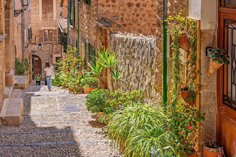 Idyllic cobblestone alley with green plants and flowers at the house walls and walking people a sunny summer day, Fornalutx, Mallorca, Spain