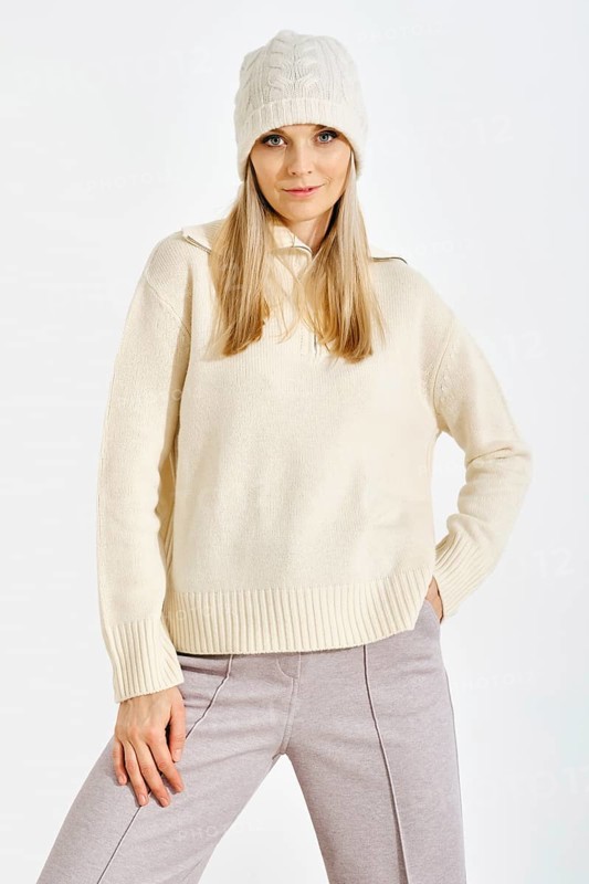 A model stands confidently in a studio while wearing a light sweater and a knitted hat. The setting features bright lighting that highlights her outfit and expression as she poses for the camera